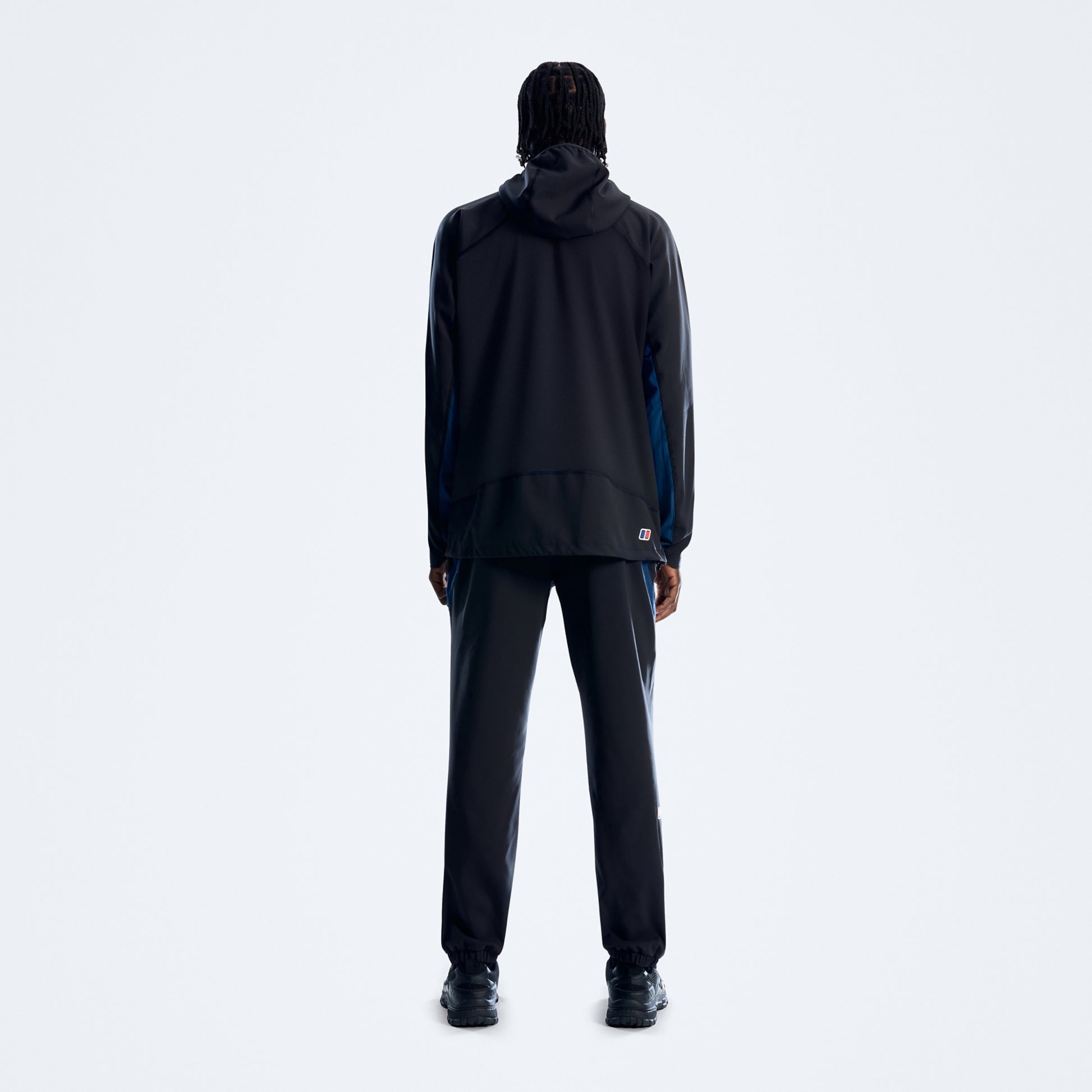 A model viewed from the back wearing a black hooded jacket with blue side panels and matching black pants, standing against a light gray studio background.