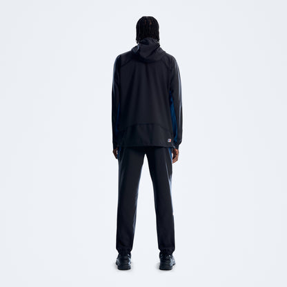 A model viewed from the back wearing a black hooded jacket with blue side panels and matching black pants, standing against a light gray studio background.