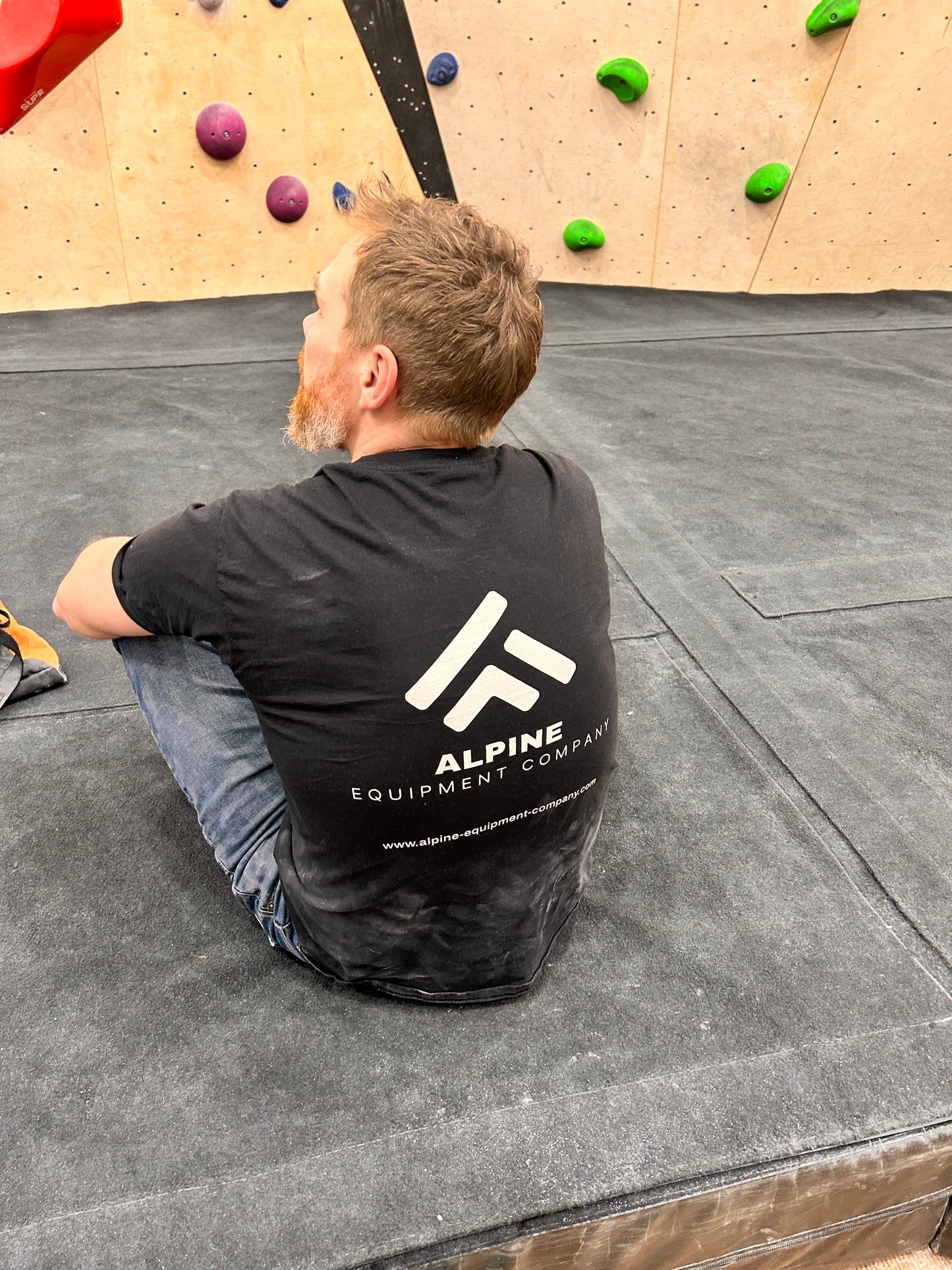 Person sitting on a climbing mat with an Alpine Equipment Company shirt on, in front of a climbing wall.