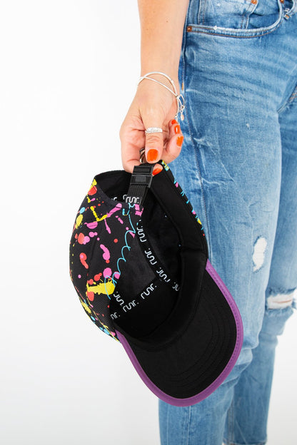 Black technical running hat with paint-splash back panel and purple brim, displayed near jeans.