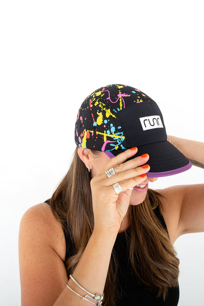 Black technical running hat with paint-splash pattern and RUNR patch, worn by woman adjusting.