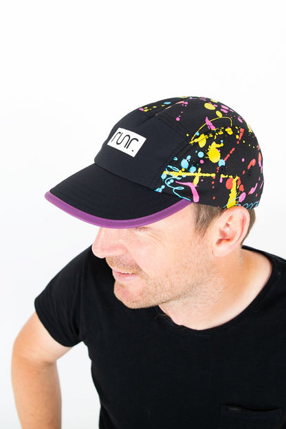 Black technical running hat with multicolor splatter print, purple brim, worn by man.
