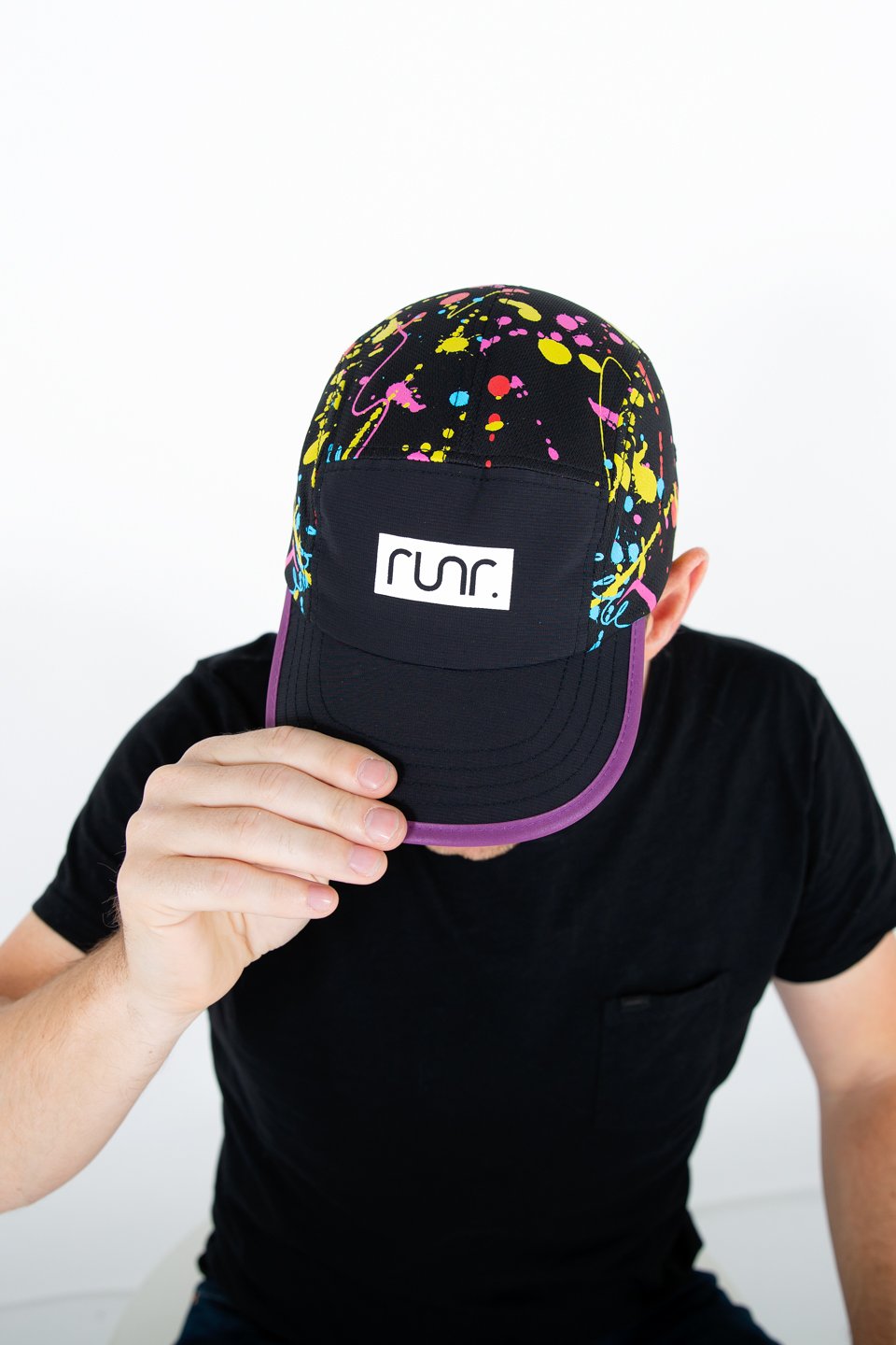 Black technical running hat with multicolor paint-splash top, purple brim, held by hand.