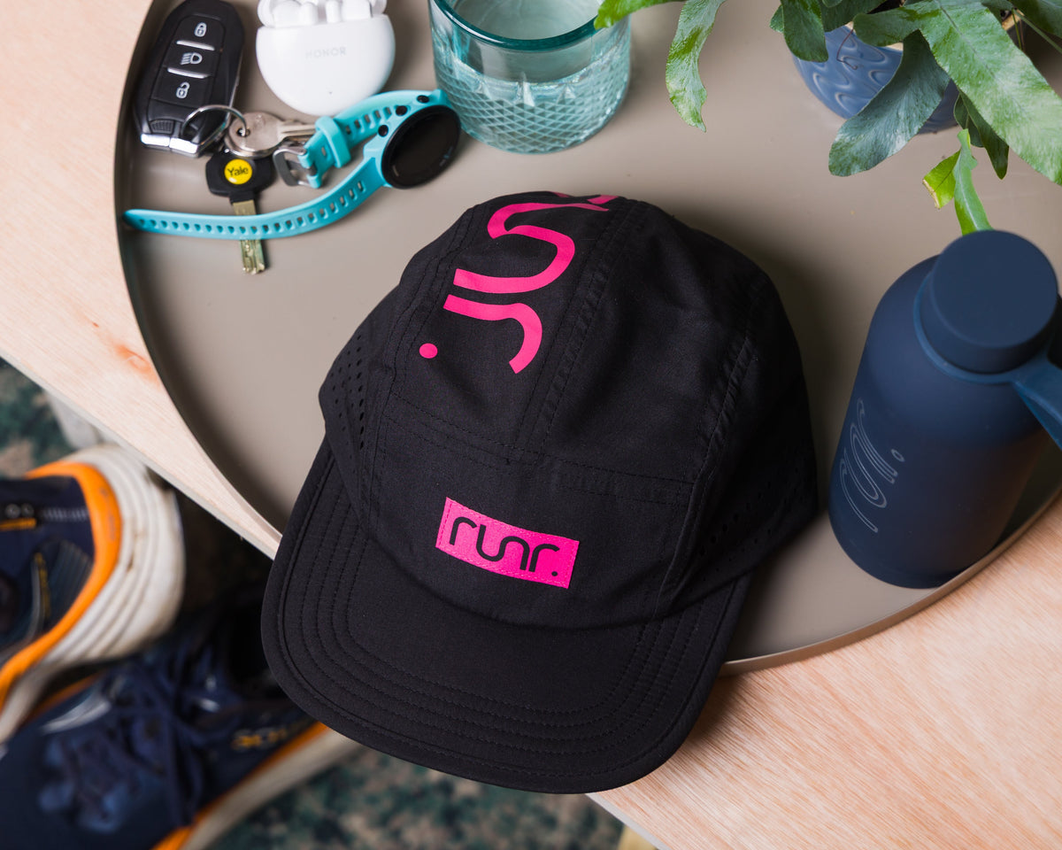 Black running cap with pink emblem on front, placed on a skateboard deck.