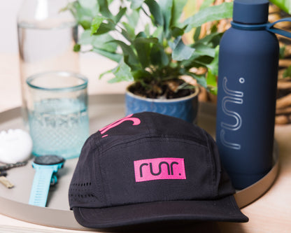 Black running cap with pink front logo, shown on a desk with bottles nearby.