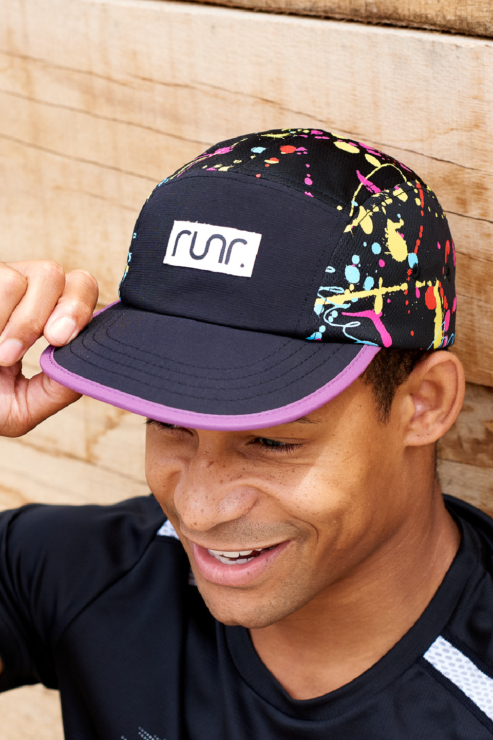 Black technical running hat with paint-splash side panel, purple underside brim, held by hand.