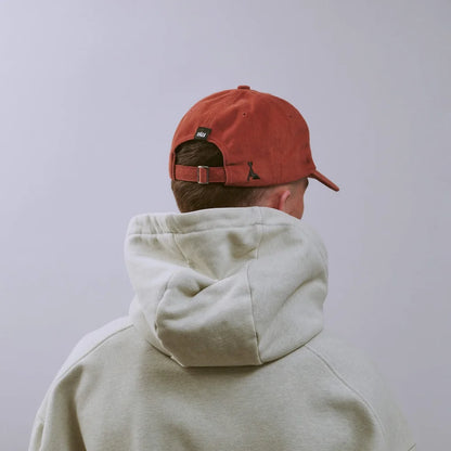 A person shown from behind wearing a rust-orange baseball cap and a light beige hooded sweatshirt against a plain light gray background.